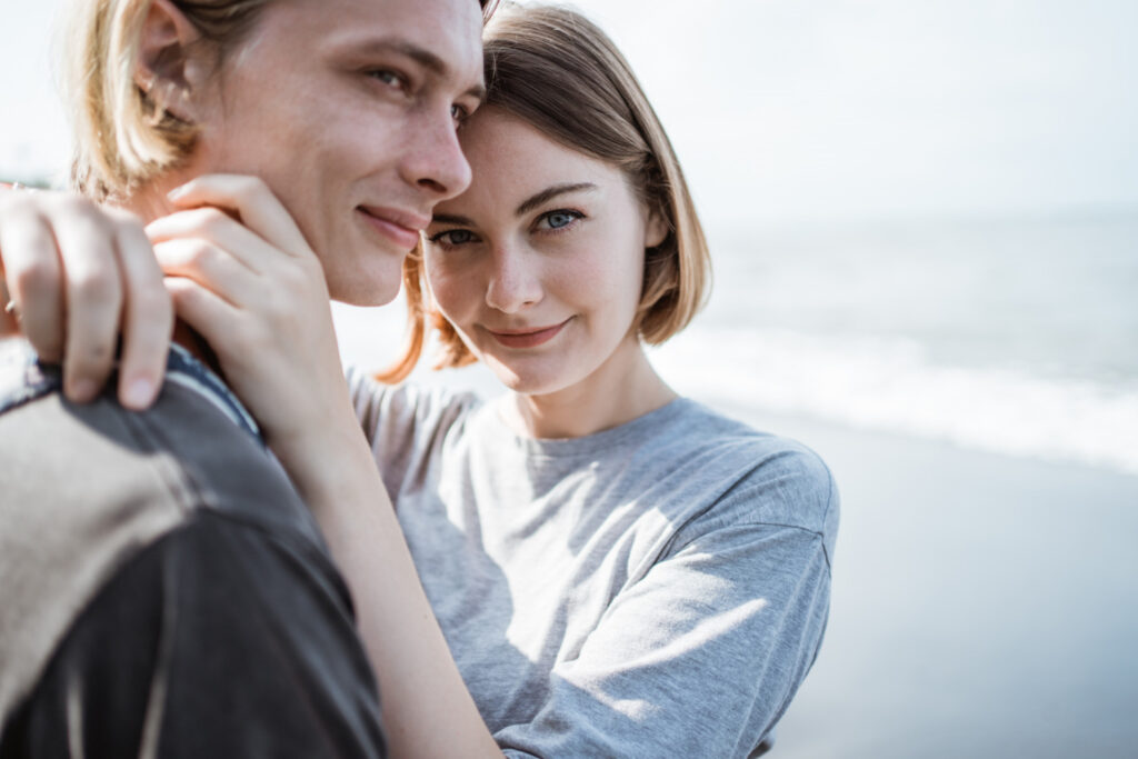 A close-up shot of a young man and woman in a gentle embrace at the beach. The woman is smiling softly at the camera with her arm around the man's neck, with the blurred ocean waves in the background.